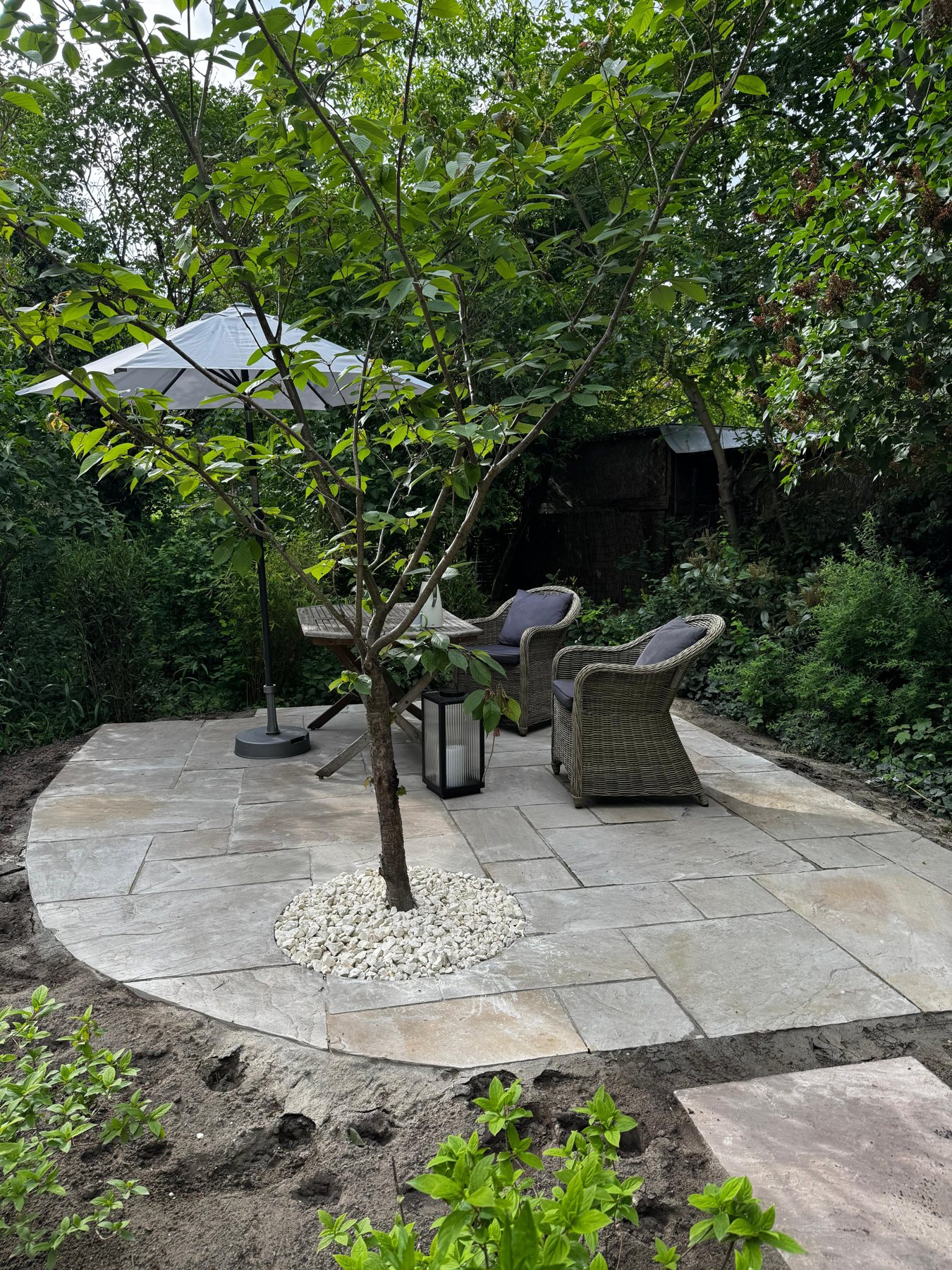 Runde Natursteinterrasse mit Baum, Sitzgruppe und Sonnenschirm im Garten