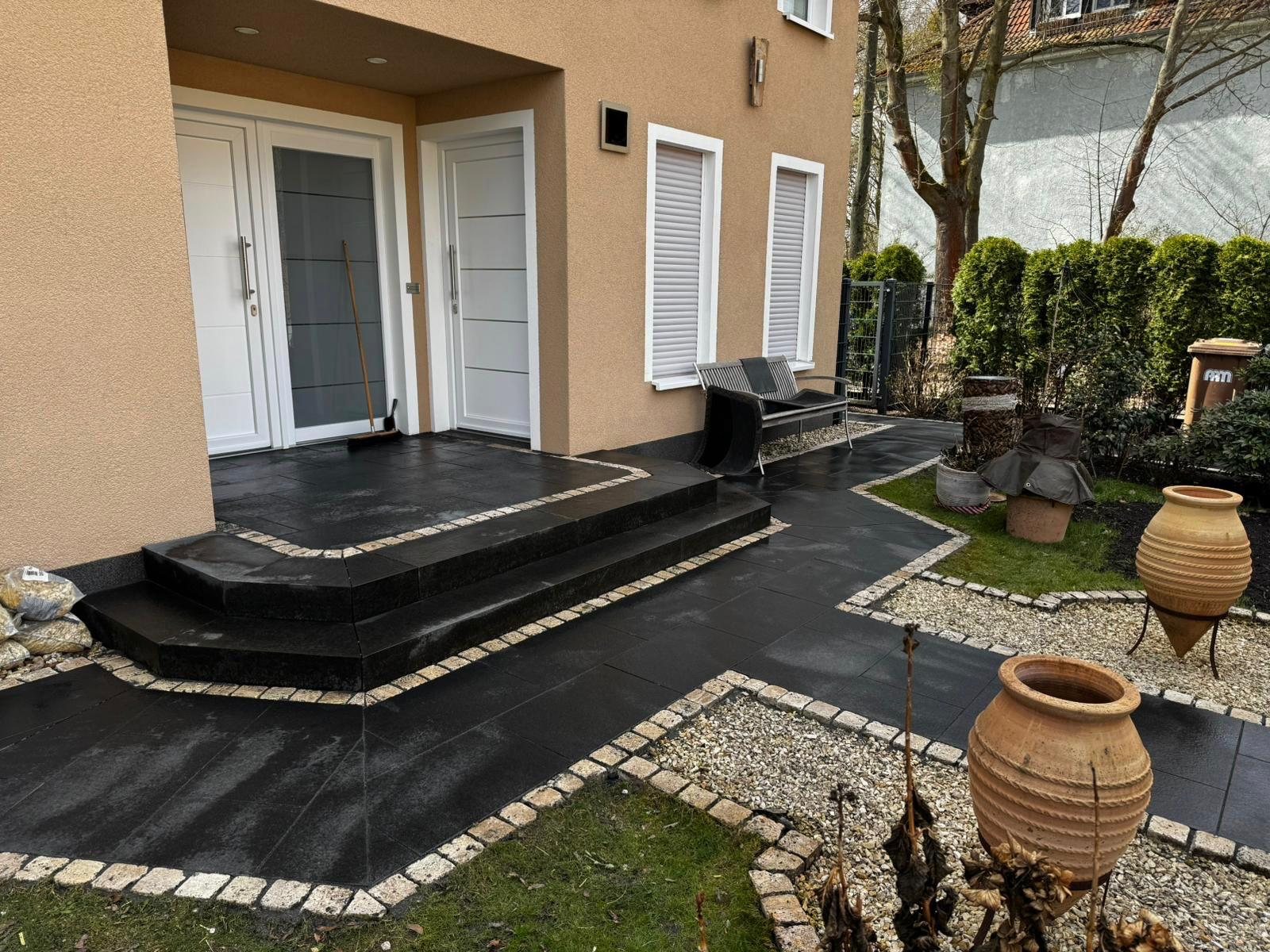 Dunkle Terrassenplatten mit heller Pflastereinfassung und Rasenstreifen vor einem Hauseingang
